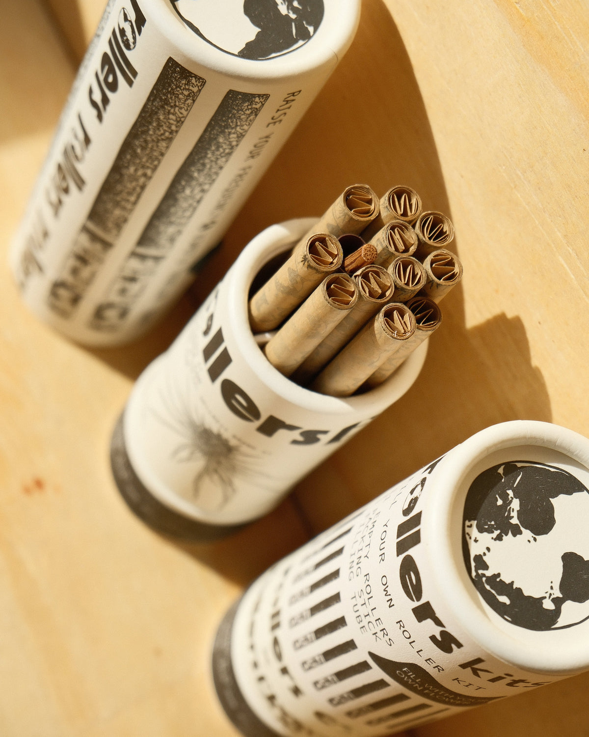 Three white cylindrical containers with black printed graphics and text, holding rolled brown sticks, arranged on a light wood surface in warm sunlight.